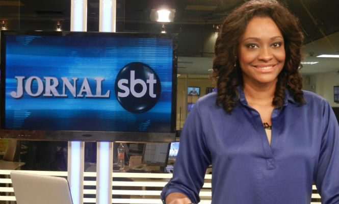 Mulher sorridente de cabelos cacheados médios, usando blusa azul, está em um estúdio de televisão ao lado de uma tela que exibe o logotipo “Jornal SBT”.