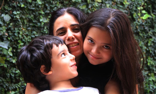 Mulher abraça duas crianças, um menino pequeno de cabelos castanhos curtos e uma menina de cabelos longos e lisos. Os três sorriem próximos uns dos outros, em frente a um muro coberto por folhagem verde.