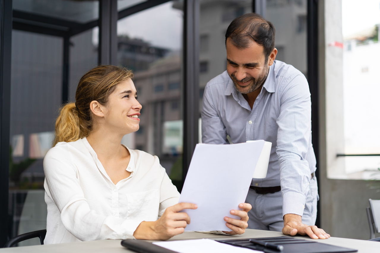 Colegas de trabalho sorridentes. O homem está em pé e a mulher sentada segurando folhas de papel.