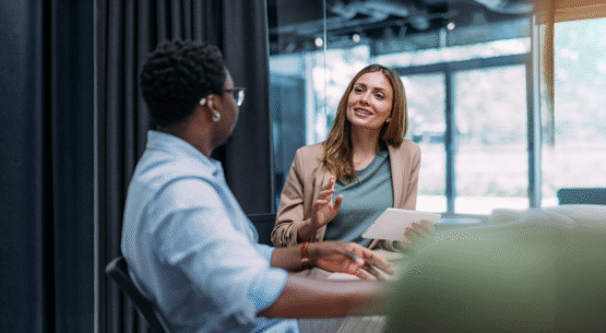 Um homem negro e uma mulher branca sentados em uma mesa de escritório conversando.